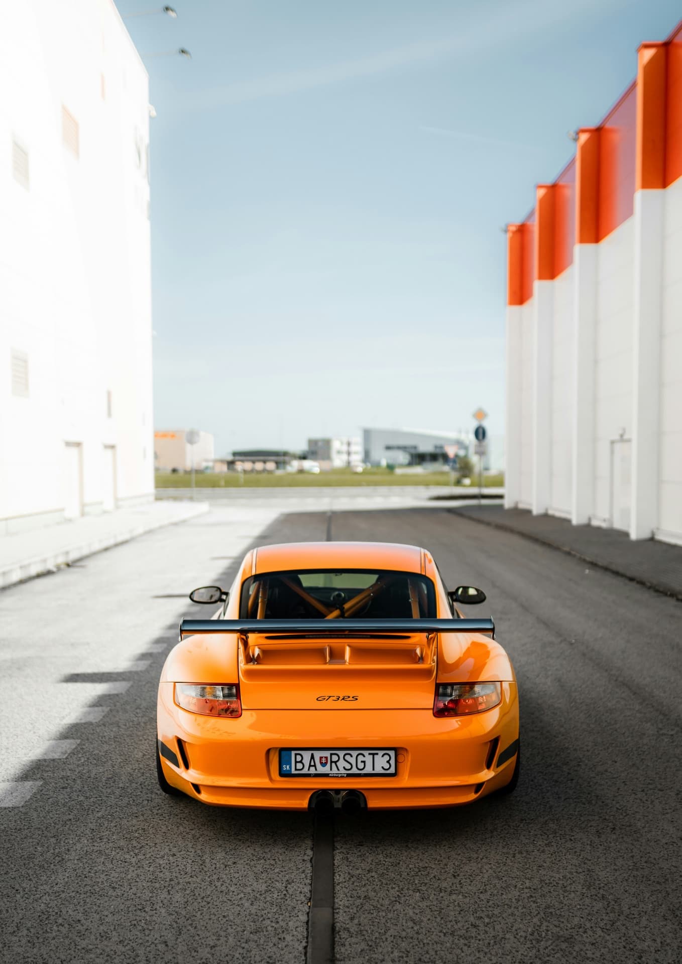 Оранжевый Porsche GT3 RS с открытым багажником припаркован на дороге между белыми зданиями